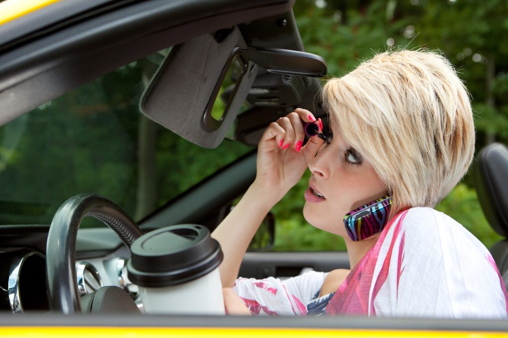 Young woman applying makeup, talking on the phone and drinking coffee while driving distracted. Seatbelt not attached.